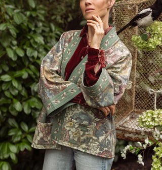 Model with hand raised to chin wears a green bell-sleeved, floral  jacket with front pockets and a bumblebee print border paired with a red velvet button-down underneath; a bird is perched behind and to the side