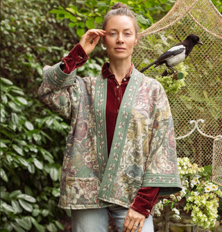 Model with hand raised to temple wears a green bell-sleeved, floral  jacket with front pockets and a bumblebee print border paired with a red velvet button-down shirt underneath; a bird is perched behind and to the side