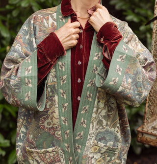 Model wears a green bell-sleeved, floral  jacket with front pockets and a bumblebee print border paired with a red velvet button-down underneath