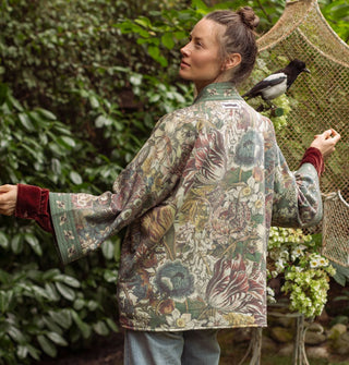 Model facing away from the camera with arms outstretched wears a green bell-sleeved, floral  jacket with front pockets and bumblebee print borders paired with a red velvet long-sleeved shirt underneath; a bird perches behind and to the side