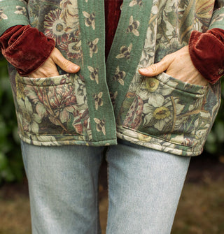 Model places hands in the front pockets of a green open-front jacket with floral print and contrasting bumblebee print border paired with a red velvet long-sleeved shirt underneath