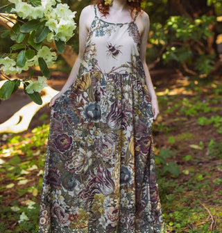 Model in a forest setting wears a lushly floral patterned slip dress with large bee illustration on the bodice