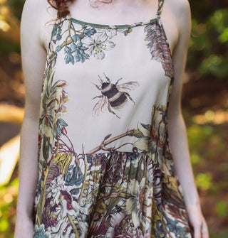 Model in a forest setting wears a lushly floral patterned slip dress with large bee illustration on the bodice