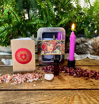 Contents of a Love Spell kit: journal with radiant heart emblem, amber vial, rose quartz, purple candle, and tin case, all staged with scattered stones and petals on a wooden tabletop against a botanical backdrop