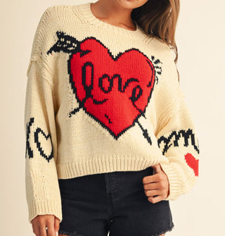 Model wears a cream-colored sweater with graphic arrow-pierced red "Love" heart design, an X and heart-shaped O on one sleeve, and "Amour" on the other sleeve