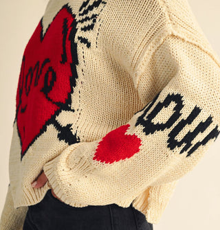 Model wears a cream-colored sweater with graphic arrow-pierced red "Love" heart design and "Amour" on the sleeve above another red heart