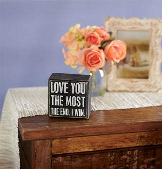 Love You the Most box sign is staged with peach-colored roses in a vase and a framed photograph on a wooden cabinet with textured runner against a purple backdrop