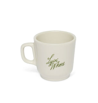 White coffee mug says "Lyin' Whore" in green script lettering on a tilt