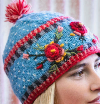 Model wears a blue wool beanie with textural, colorful floral embroidery and a pink pom pom on top
