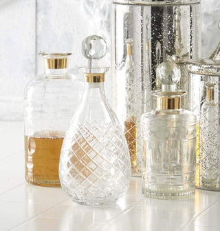 Faceted glass decanters with gold band trim on a tiled surface with a speckled reflective ice bucket; one decanter is partially filled with liquid