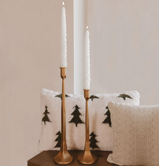 Set of two lit white evergreen tree-shaped taper candles in copper-colored holders sit on a wooden bench accented with seasonal pillows