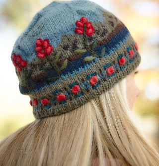 Model looking downward wears a colorful wool beanie with a mountain scene over-stitched with raised floral embroidery