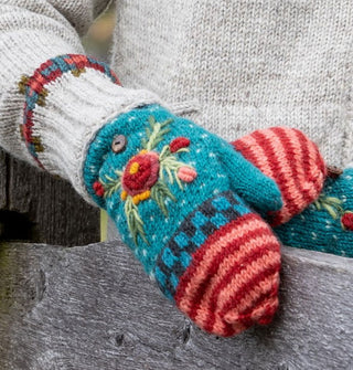 Model leaning on a rusting fence wears a pair of colorful knit mittens with raised floral embroidery