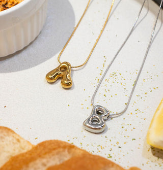 Gold "A" and silver "B" necklaces with bubble letter pendants and cable chains rest on a surface staged with bread, butter, and crème brûlée