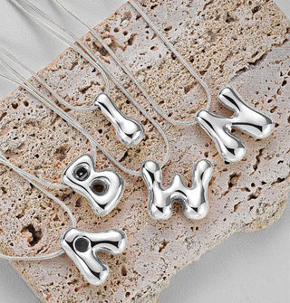 Grouping of silver necklaces with cable chains and bubble letter pendants rest on a piece of porous stone