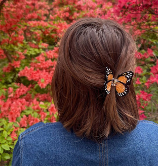 Model wears an orange monarch butterfly hair clip in the back of a partially swept-back style against a backdrop of reddish-orange flowers