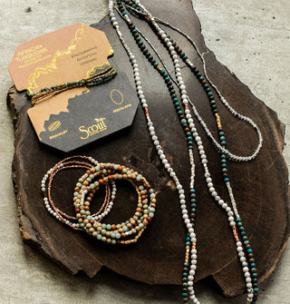 Collection of stone bead jewelry arranged on a wood platform, one of which is wrapped around a product card accented in gold foil