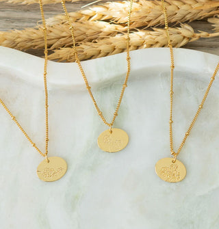 Three gold necklaces with flower designed etched into oval pendants ion satellite chains rest on a marble dish next to stalks of wheat