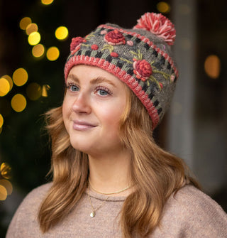 Smiling model wears a colorful knit beanie with textural floral embroidery, border details, and a two-tone pom pom at the top