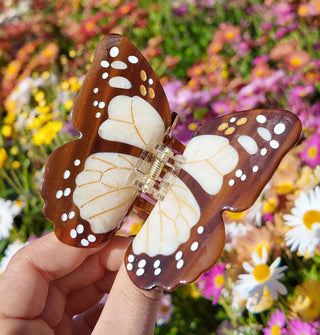 Model's hand holds a brown and white monarch butterfly hair clip against a backdrop of colorful flowers