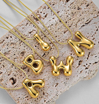 Grouping of gold necklaces with cable chains and bubble letter pendants rest on a piece of porous stone
