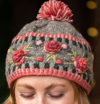 Model wears a colorful knit beanie with textural floral embroidery, border details, and a two-tone pom pom at the top