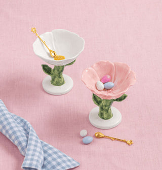 Hand-painted ceramic flower pedestal dishes with accompanying gold spoons are staged with pastel egg candies and a blue and white gingham tea towel on a pink surface