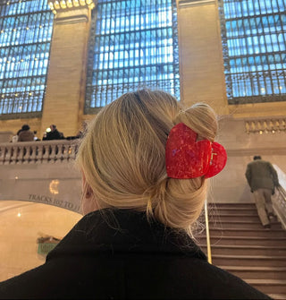 Model standing in a train station wears a red, heart-shaped hair clip in a twist style