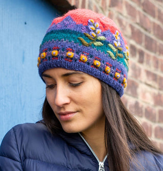 Model looking downward wears a colorful wool beanie with a mountain sunset scene over-stitched with raised floral embroidery
