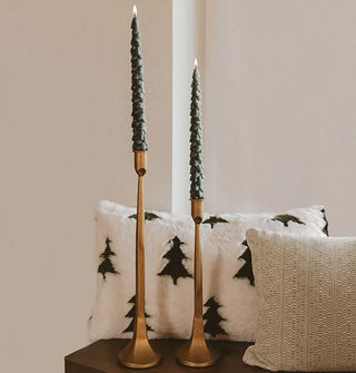 Set of two lit evergreen tree-shaped taper candles in copper-colored holders sit on a wooden bench accented with seasonal pillows