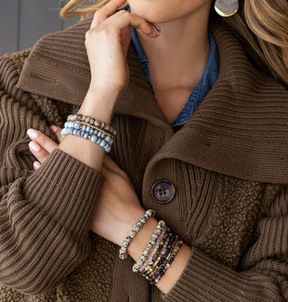 Model wears stacks of stone bead bracelets on each wrist with a brown sweater