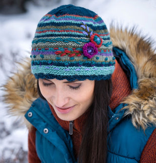 Model in fur-lined puffy blue vest wears a striped wool beanie with textural embroidered flower accents against a snowy backdrop
