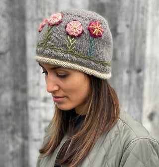 Model looking downward wears a gray wool beanie with beige ribbed band and colorful embroidered flowers all across its middle