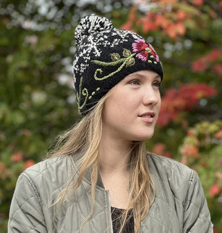 Model wears a black beanie with pink, purple, and green floral embroidery around the band, white snowflake pattern around the crown, and a black and white pom pom at the top