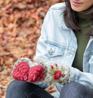 Model sitting against a backdrop of autumn leaves wears a pair of colorful knit mittens with raised floral embroidery