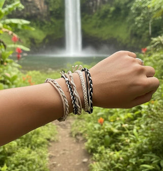 Model's arm held up against a tropical waterfall backdrop wears several woven hair elastics on wrist in a neutral palette