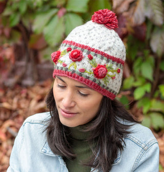 Model wears a white wool beanie with textural, colorful floral embroidery and a pink pom pom on top
