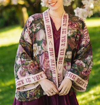Smiling model wears a cropped silken robe with lush pink and purple floral print and contrasting trim that says, "Wish with me, the best is yet to be"