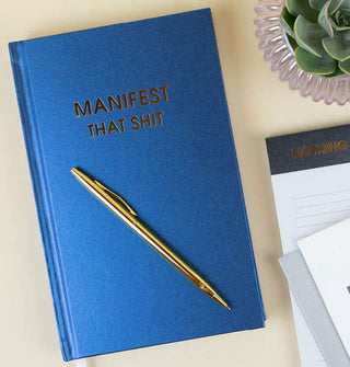Blue hardcover Manifest That Shit journal with gold pen atop sits next to a potted succulent and lined writing pad