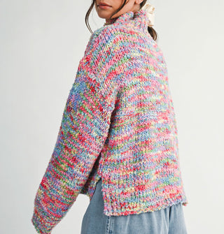 Model turning away from the camera wears a multicolored, oversized  turtleneck sweater with bottom side slit
