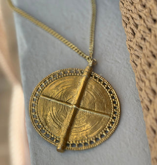 Large round gold-toned medallion with lacy edge and central "t" or cross formed by two perpendicular ridges is attached to a thin curb chain