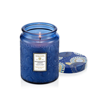 Lit single-wick Mediterranean Lemon Voluspa candle in embossed blue glass with a matching floral-patterned tin lid removed and set to the side