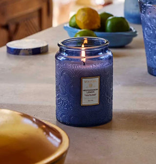 Lit single-wick Mediterranean Lemon Voluspa candle in a blue embossed glass jar rests on a countertop with other blue glass, citrus fruit, and its tin lid staged in the background and foreground