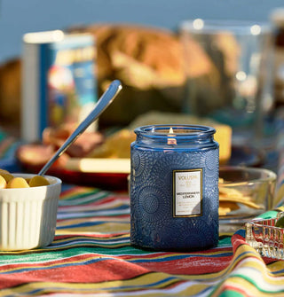 A lit Mediterranean Lemon candle by Voluspa in an embossed blue glass vessel is staged on a colorful striped tablecloth next to a dish of olives