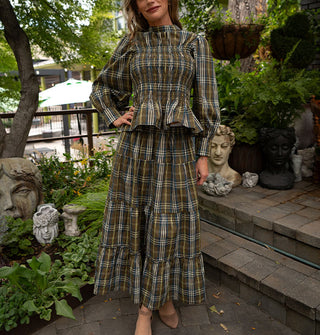 Model posing with one hand on hip wears a plaid blouse and skirt set with ruffled tiers in an olive green, yellow, white, and blue palette