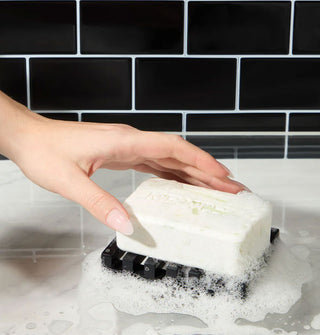 Model's hand touches a lathered bar of soap on a black ridged tray against a black tiled backdrop