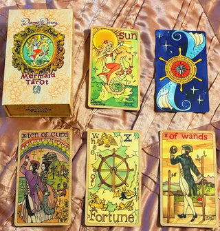 Dame Darcy Mermaid Tarot box with five sample cards from the deck: The Sun, a card turned upside-down to show its back, Ten of Cups, Wheel of Fortune, and Two of Wands, all resting atop rose gold satin