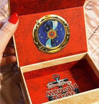 Model's hand holds open the Dame Darcy Mermaid Tarot card box, which features red patterned partial lining and a ship's wheel insignia