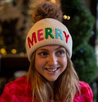 Smiling model wears a white knit beanie that says, "Merry" on the folded cuff in multicolored lettering and has a brown faux fur pompom on top
