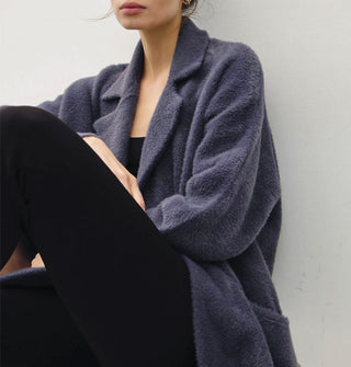 Seated model wears a long, dark muted blue fuzzy coat/cardigan with notched collar and pocket
